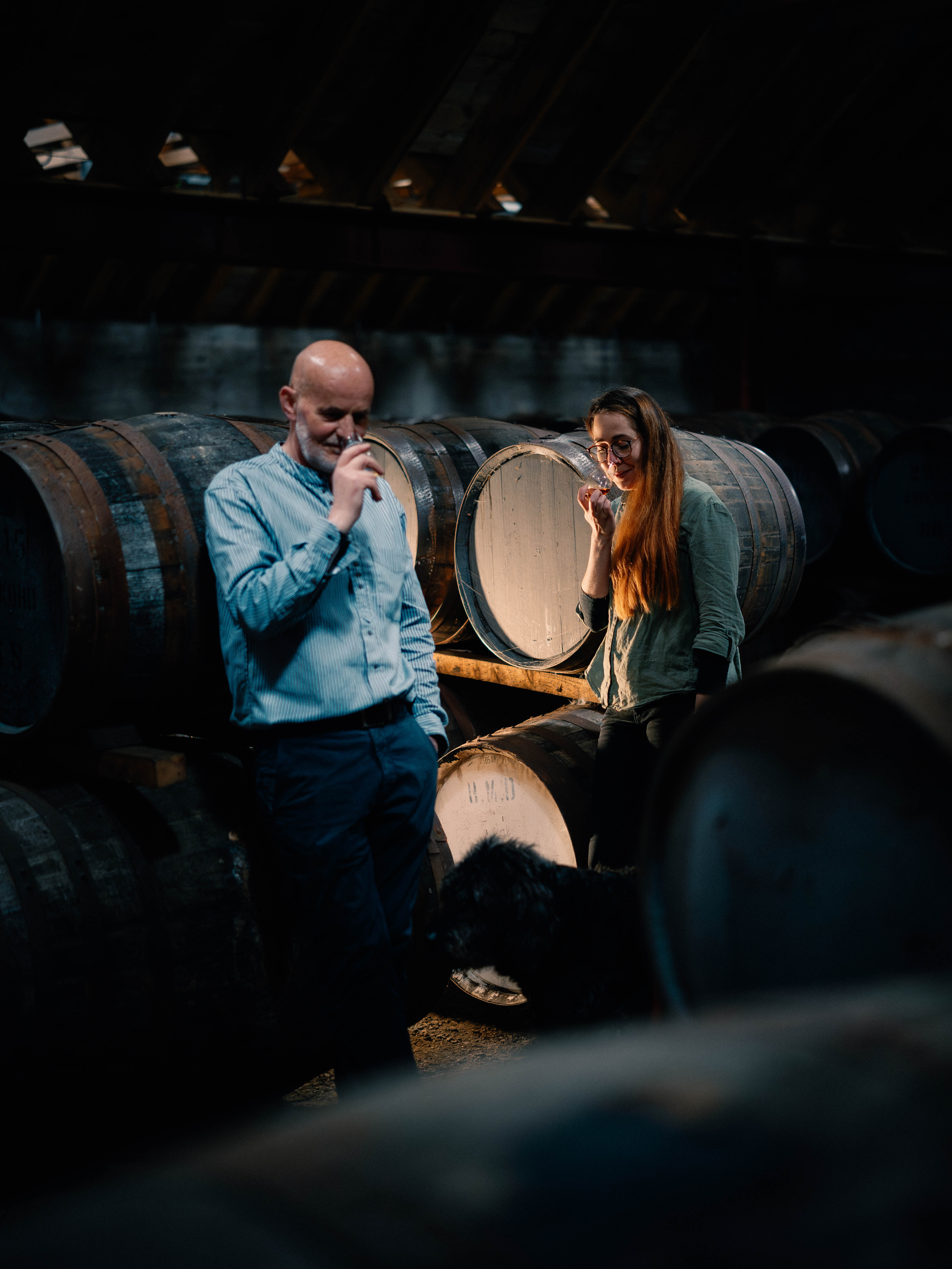 A man and a woman hold up small glasses of whisky to their noses. They are surrounded by a number of wooden casks